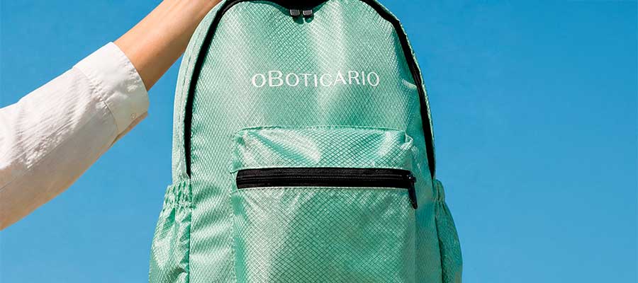 Mochila dobrável personalizada verde-água O Boticário segurada pela alça contra céu azul, mostrando bolso frontal e zíper preto